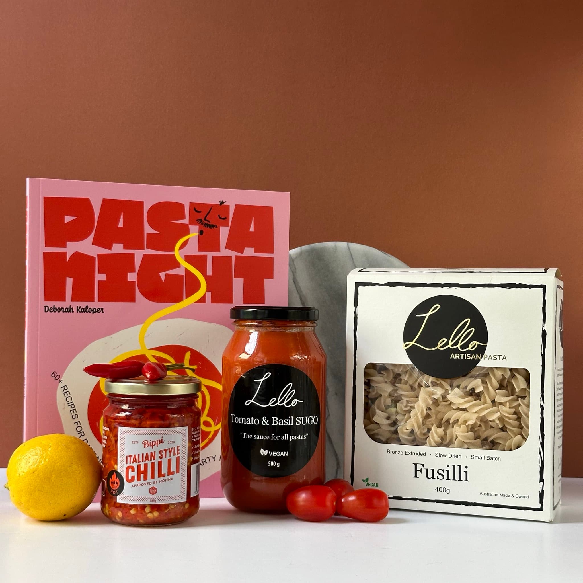 The items include a pink cookbook titled "Pasta Night" by Deborah Kaloper, a jar of Bippi Italian Style Chilli, a jar of Lello Tomato & Basil Sugo, a box of Lello Fusilli pasta, fresh cherry tomatoes, and a lemon. A round marble plate is partially visible in the background.